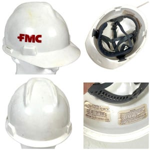 May include: White hard hat with the red letters "FMC" on the front. The interior features a suspension system and adjustable head straps. The hard hat has a brim and is designed for safety.