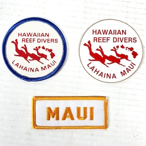 Può includere: Tre toppe: due circolari e una rettangolare. Le toppe circolari riportano "HAWAIIAN REEF DIVERS LAHAINA MAUI" con subacquei rossi e una mappa. Una ha un bordo blu, l'altra un bordo bianco. La toppa rettangolare riporta "MAUI" in oro.