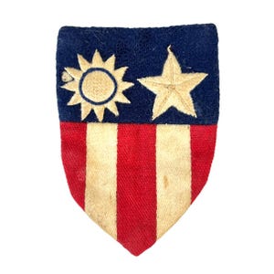 May include: A vintage shield-shaped patch with a blue top section featuring a sun and a star, and a red and white striped bottom. The patch is made of fabric and has a worn appearance.