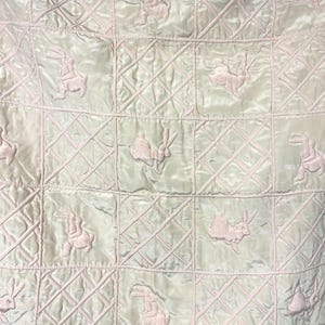May include: A light green quilted blanket with a repeating pattern of pink rabbits. The blanket is divided into square sections with a diamond pattern stitched into each square. The rabbits are embroidered in a soft pink color.