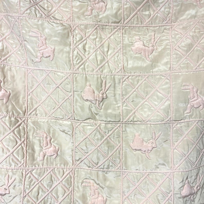 May include: A light green quilted blanket with a repeating pattern of pink rabbits. The blanket is divided into square sections with a diamond pattern stitched into each square. The rabbits are embroidered in a soft pink color.