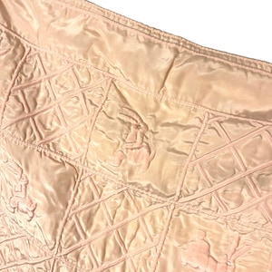 May include: A light pink quilted fabric with a repeating pattern of diamond shapes and bunny rabbit figures. The fabric has a satin sheen and a raised, textured design. The bunnies are in a light pink color.