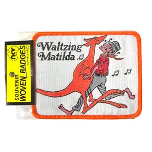 Puede incluir: Un parche de recuerdo rectangular con un borde naranja. El parche presenta una ilustración de dibujos animados de una persona bailando con un canguro, con el texto "Waltzing Matilda". Se adjunta una etiqueta amarilla con texto.