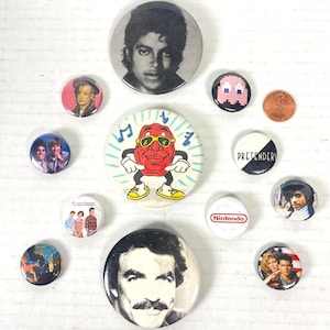 May include: A selection of vintage pin badges showcasing pop culture figures. Badges include images of Michael Jackson, Boy George, Prince, and a Pac-Man character. Other badges feature the Raisin Bran mascot and the band The Pretenders.