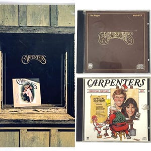 May include: A collection of Carpenters CDs. One CD case is brown with the text "The Singles 1969-1973". Another CD case is white with the text "Christmas Portrait". The third CD case is white with the text "Carpenters".