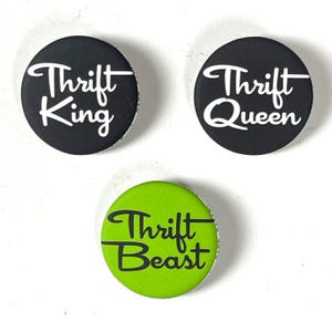 Może przedstawiać: Trzy okrągłe guziki z białym tekstem na czarnym tle i czarnym tekstem na zielonym tle. Guziki mają napis "Thrift King", "Thrift Queen" i "Thrift Beast".