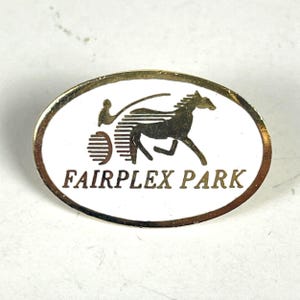 May include: A white oval pin with a gold border and a gold silhouette of a horse and driver. The text "FAIRPLEX PARK" is printed in gold below the image.