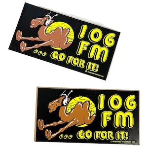 May include: Two rectangular stickers featuring a cartoon camel wearing headphones, set against a yellow sun. The text "106 FM GO FOR IT!" is in yellow. The stickers are on a black background.
