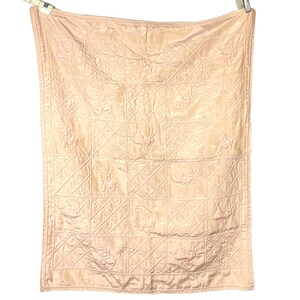 May include: A light pink, quilted blanket with a grid pattern. The blanket features embroidered floral and bunny designs within each square. The blanket is hanging from a clothesline, showcasing its texture and design.