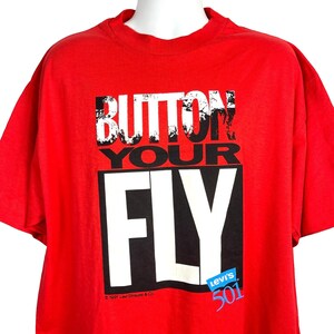Puede incluir: Camiseta roja de manga corta con el texto "Button Your Fly" en letras blancas y negras. El texto "Levi's 501" está en un cuadro azul con letras blancas. El texto de copyright "© 1991 Levi Strauss & Co." está en la parte inferior de la camiseta.