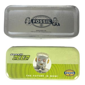 May include: Two rectangular Fossil tins. The top tin is silver with the Fossil logo and vintage phone illustrations. The bottom tin is green with a robot illustration, the text "Fossil 2002", and "THE FUTURE IS NOW!"