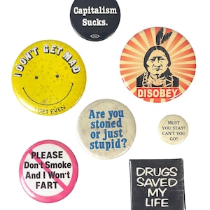 May include: A collection of vintage buttons with various designs and text. Buttons include a yellow smiley face with "I DON'T GET MAD GET EVEN", a black button with "Capitalism Sucks", and a button with "DISOBEY".