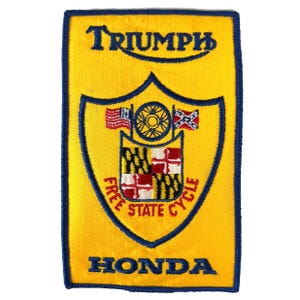 May include: Yellow embroidered patch with a blue border. The patch features the word "TRIUMPH" at the top and "HONDA" at the bottom. A shield design in the center includes flags, a wheel, and the words "FREE STATE CYCLE".