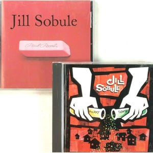 May include: Two compact discs with different album covers. The first CD has a pink background with the text "Jill Sobule" and a pink eraser with the text "Pink Pearl". The second CD has a red, black, and white abstract design with the text "Jill Sobule" and the words "Happy Town" on two green and yellow objects.