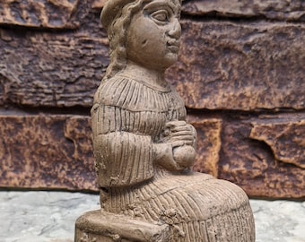 May include: A brown stone sculpture of a seated Sumerian woman holding a small vessel. The sculpture is 7.87 inches tall and is made of aged stone.