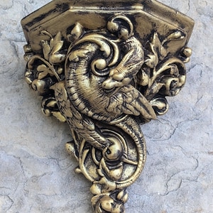 May include: A gold-toned wall shelf with a decorative design of a dragon with wings and a tail. The shelf is made of resin and has a distressed finish. The shelf is 7 inches tall and 2 inches wide.  Gargoyle Satyr dragon 7" 2 Tone Gold Aged
