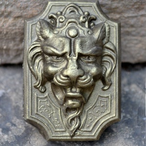 May include: A bronze-colored gargoyle bust sculpture with a detailed face and a beard. The bust is mounted on a rectangular plaque with ornate designs.