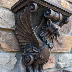 May include: A decorative wall bracket made of dark brown wood, carved in the shape of a mythical griffin with outstretched wings. The griffin has a detailed design with intricate carvings and a textured surface.