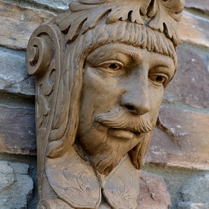 May include: A brown stone wall bracket corbel featuring a sculpted man's head with a crown and scrollwork. The corbel is 12 inches tall and has an aged stone finish. "French Man wall bracket corbel 12" Aged Stone" is written below the corbel.