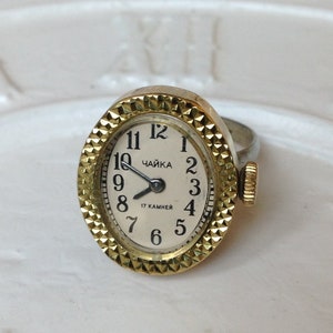 May include: A gold-tone ring watch with a white face and black numbers. The watch face reads "ЧАЙКА 17 КАМНЕЙ".