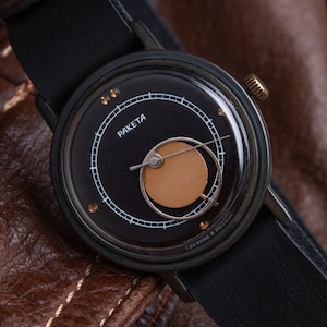 May include: A black wristwatch with a leather strap, featuring a dark face with gold accents and the word "Raketa". The watch is set against a brown leather jacket, showcasing its design and style.