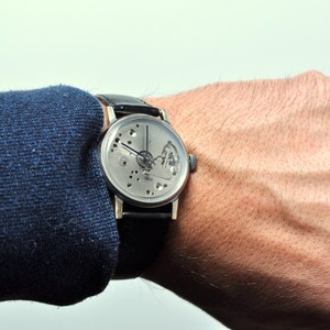 May include: A silver wristwatch with a skeletonized dial and black leather band. The watch is worn on a person's wrist.