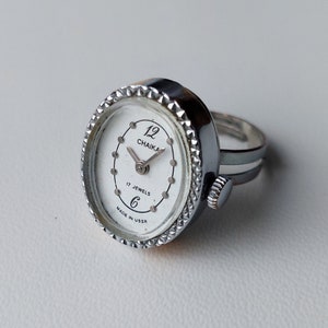 Może przedstawiać: Srebrny pierścionek z zegarkiem o białej tarczy. Tarcza zegarka ma czarne cyfry i wskazówki, a także napis "12 CHAIKA 17 JEWELS MADE IN USSR".
