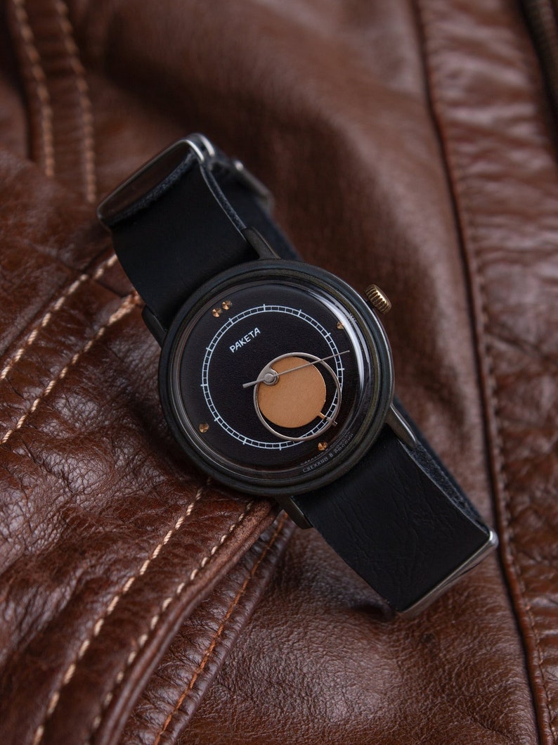 May include: A black wristwatch with a leather strap, featuring a dark face with gold accents and the word "Raketa". The watch is set against a brown leather jacket, showcasing its design and style.