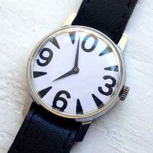 May include: A black leather wristwatch with a white face and black numbers. The numbers are arranged in a unique pattern, with the 12 at the top, the 3 at the right, the 6 at the bottom, and the 9 at the left. The watch has a silver case and a black leather band.