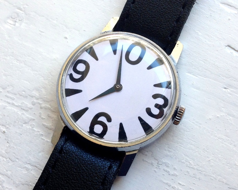 May include: A black leather wristwatch with a white face and black numbers. The numbers are arranged in a unique pattern, with the 12 at the top, the 3 at the right, the 6 at the bottom, and the 9 at the left. The watch has a silver case and a black leather band.