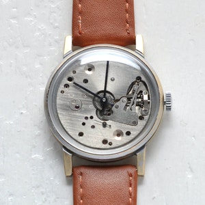 May include: A gold-tone wristwatch with a brown leather band. The watch face is clear and shows the inner workings of the watch.