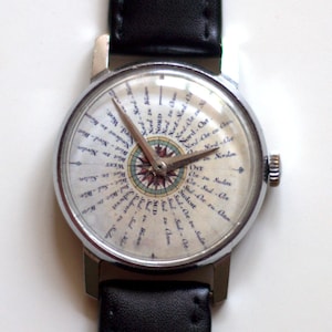 May include: A silver wristwatch with a black leather band. The watch face has a compass design with the words "Nord", "Sud", "Ost", and "West" written around the compass.
