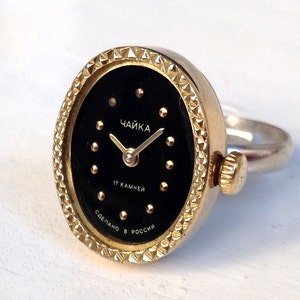 May include: A gold-tone ring watch with a black face. The watch face has gold-tone hands and hour markers. The watch face also has the text "Чайка" and "17 камней" in Cyrillic script. The watch band is silver-tone.