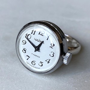 Può includere: Anello d'argento con quadrante bianco, numeri neri e lancette nere. Il quadrante presenta la scritta "ЧАЙКА" e il testo "17 KAMHEЙ" e "СДЕЛАНО В РОССИИ".