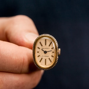 May include: A gold-toned oval wristwatch with a white face and black hands. The watch face has a black and white patterned design and the text "Romantica 17 Jewels Swiss Made" printed on it.
