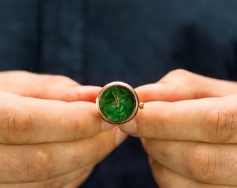 May include: A round, green, and gold watch with a white face and black hands. The watch is being held in the palm of a hand.