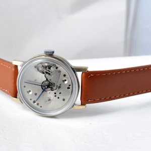May include: A silver skeleton watch with a brown leather band. The watch face is open, revealing the inner workings of the watch.