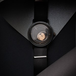 May include: A black wristwatch with a leather strap and a round face. The watch face has a dark background with a lighter circular design. The watch strap is secured with a silver clasp. The word "PARETA" is printed on the watch face.