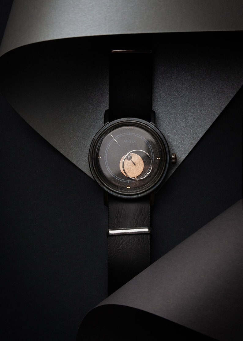 May include: A black wristwatch with a leather strap and a round face. The watch face has a dark background with a lighter circular design. The watch strap is secured with a silver clasp. The word "PARETA" is printed on the watch face.