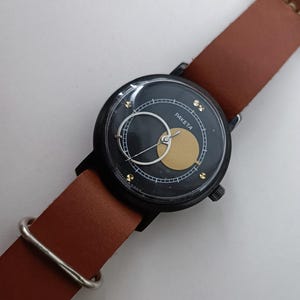 May include: A black Paketa wristwatch with a brown leather strap. The watch face has a gold circle and a white crescent. The strap is approximately 2 cm wide.