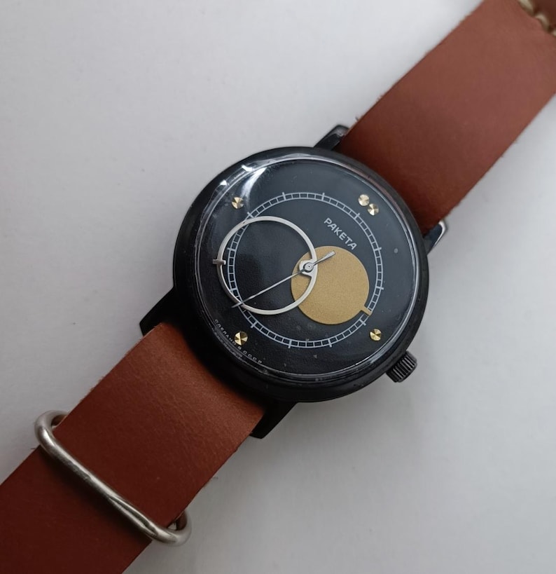 May include: A black Paketa wristwatch with a brown leather strap. The watch face has a gold circle and a white crescent. The strap is approximately 2 cm wide.