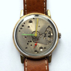May include: A gold-tone wristwatch with a brown leather band. The watch face is open, revealing the inner workings of the watch.