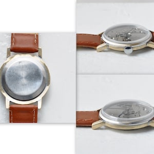May include: A gold-tone wristwatch with a brown leather band. The watch has a clear back that reveals the inner workings of the watch.