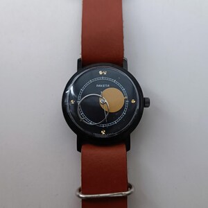 May include: A black Raketa watch with a brown leather strap. The watch face has a black background with a gold circle and a silver hand. The watch has a black case and a brown leather strap.