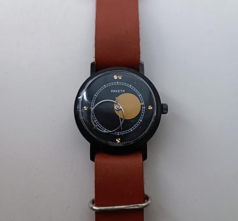 May include: A black Raketa watch with a brown leather strap. The watch face has a black background with a gold circle and a silver hand. The watch has a black case and a brown leather strap.