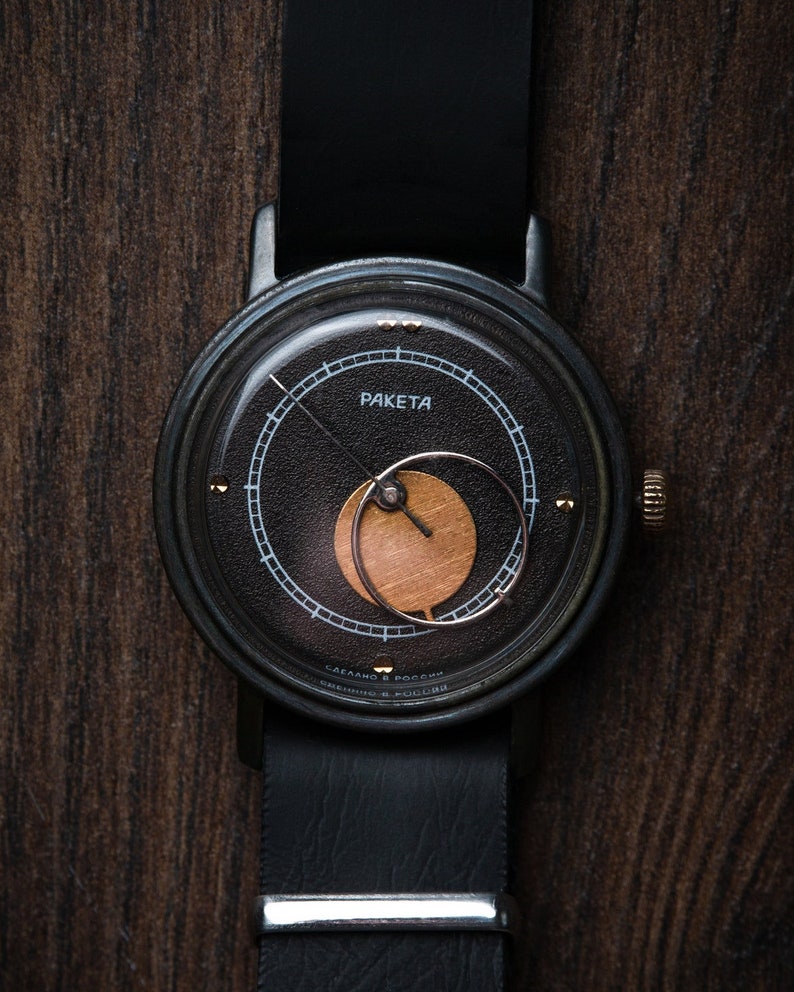 May include: A black Raketa wristwatch with a black leather strap. The watch face is black with gold and white detailing, including the word "Raketa". The watch has a gold-coloured crown and a silver strap clasp. The watch is set against a dark wood grain background.