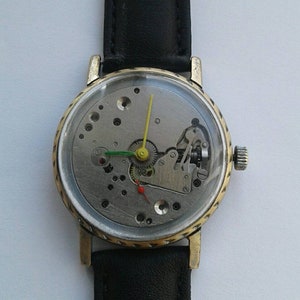 May include: A gold-toned wristwatch with a black leather band. The watch face is clear and shows the inner workings of the watch mechanism.