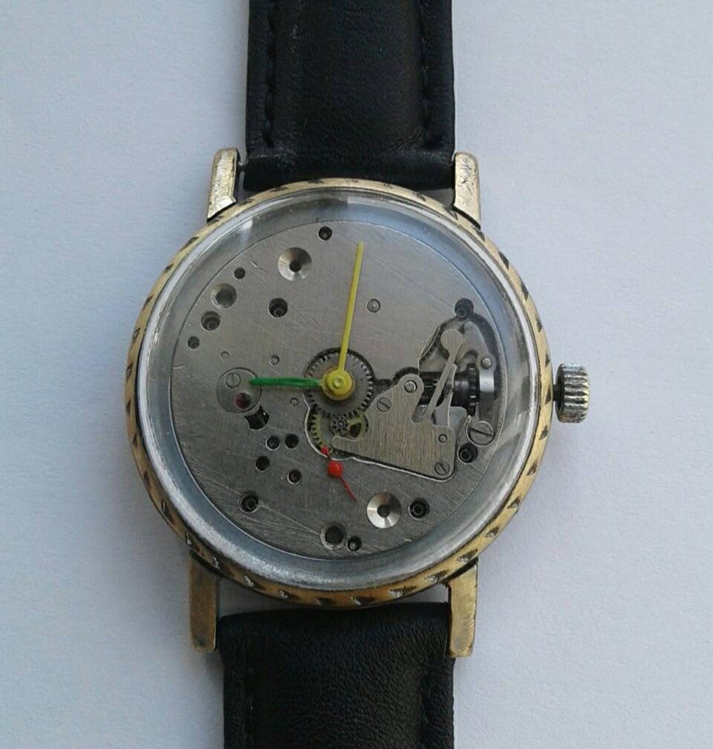 May include: A gold-toned wristwatch with a black leather band. The watch face is clear and shows the inner workings of the watch mechanism.