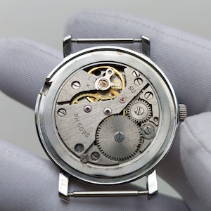 May include: Detailed view of a vintage watch mechanism, revealing the complex arrangement of gears, springs, and jewels. The silver-coloured movement is set within a round case with strap lugs. The internal workings of the watch are exposed, emphasizing its mechanical nature.