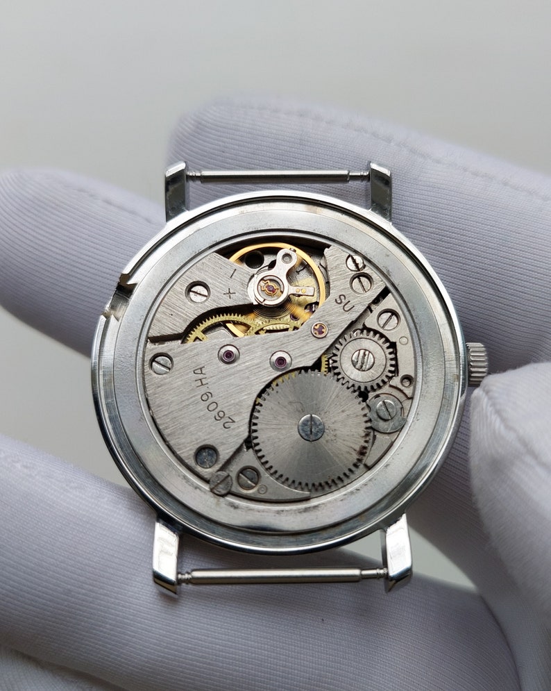 May include: Detailed view of a vintage watch mechanism, revealing the complex arrangement of gears, springs, and jewels. The silver-coloured movement is set within a round case with strap lugs. The internal workings of the watch are exposed, emphasizing its mechanical nature.